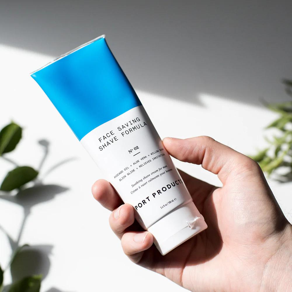 A hand holds the Port Products Face Saving Shave Formula in a blue and white tube against a white background with green plant leaves and shadows.