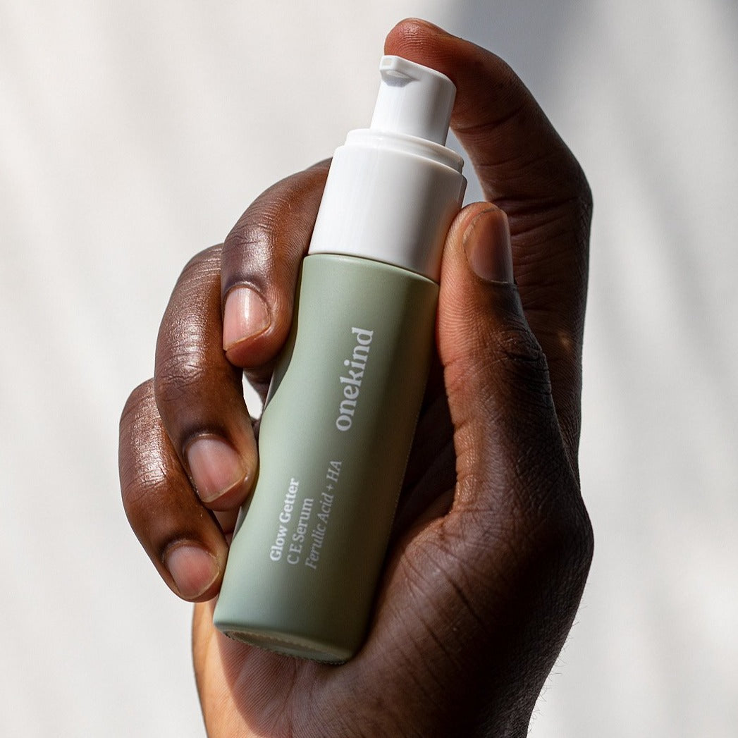 A hand holding the Glow Getter C E Serum in a sage green glass bottle with a white pump cap against a white background with shadows.