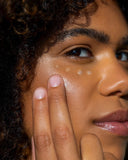 A side profile of a person with glowing skin looking toward the camera with 4 drops of the Pep Squad gel dabbed under their eye with two fingers pressed to the cheek, ready to spread the gel.