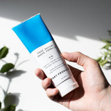 Port Products Face Saving Shave Formula white and blue tube in man's hand against a white background  with shadows and plants.