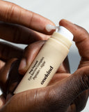 A hand holds a Pep Squad Eye Revive Complex in a light tan tube with a white pump top and is pumping the white-colored gel onto the fingertip of the other hand.