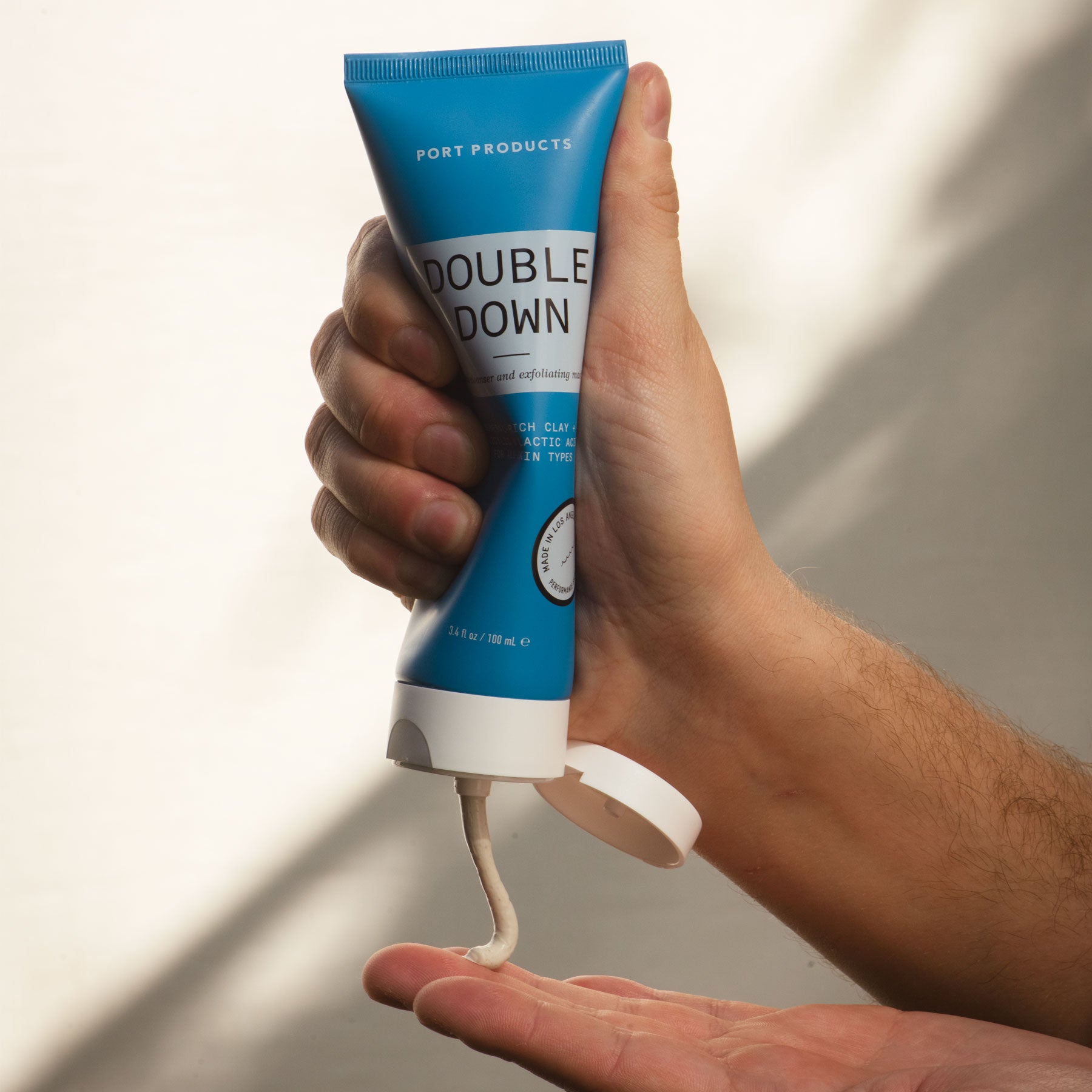 Double Down Face Cleanser and Exfoliating Mask blue tube in man's hand. One hand squeezing tube and gray cream into other hand