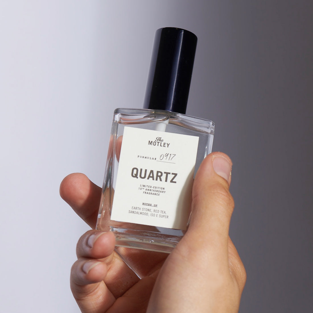 A hand holds The Motley Quartz Fragrance in a clear glass bottle with clear liquid and a black pump cap against a gray background.