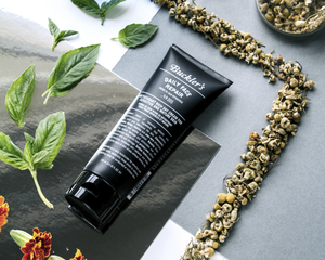 A Buckler's Daily Face Repair in a black tube with white writing lays on a counter surrounded by basil leaves and chamomile