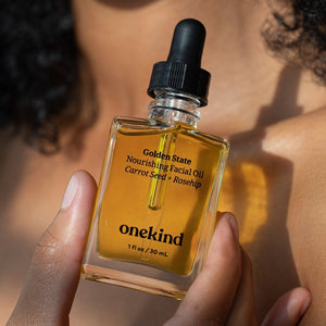 A hand holds the Golden State Nourishing Facial Oil in a clear jar that shows the golden oil with a black dropper top in the sunlight in front of their shoulder and neck.
