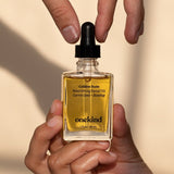 A Golden State Nourishing Facial Oil in a clear jar that shows the golden oil with a black dropper top stands against a pale brown background.