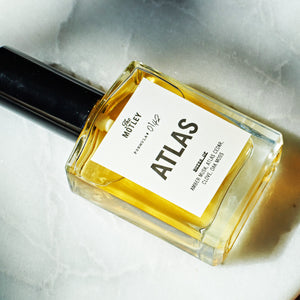 The Motley Atlas Fragrance in a glass bottle with gold liquid and a black pump cap rests on a marble counter with shadows.