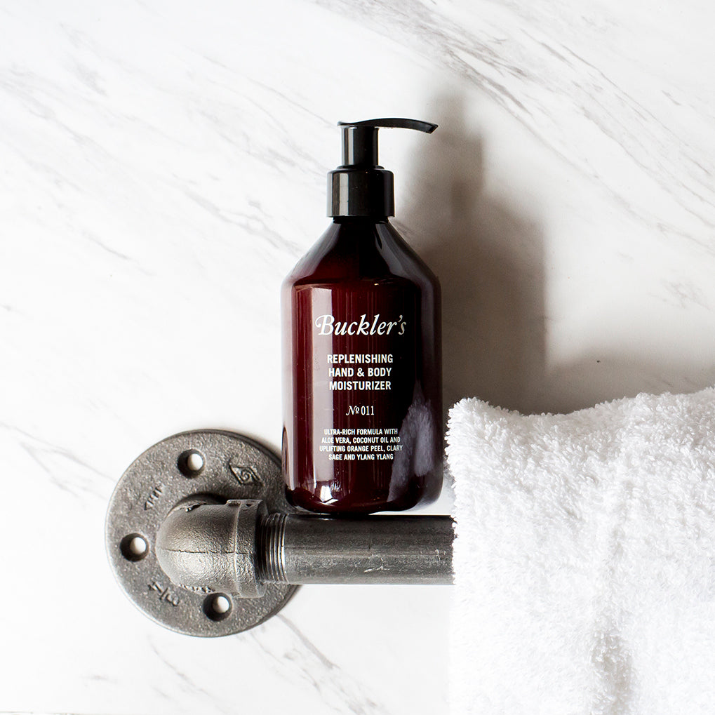 The Buckler's Replenishing Hand & Body Moisturizer in a red wine-colored bottle with a black pump top stands on a silver bathroom handle bar next to a white towel against a white and gray marble background.