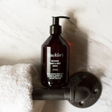 The Buckler's Reviving Hand & Body Wash in a red wine-colored bottle with a black pump top stands on a silver bathroom handle bar next to a white towel against a white and gray marble background.