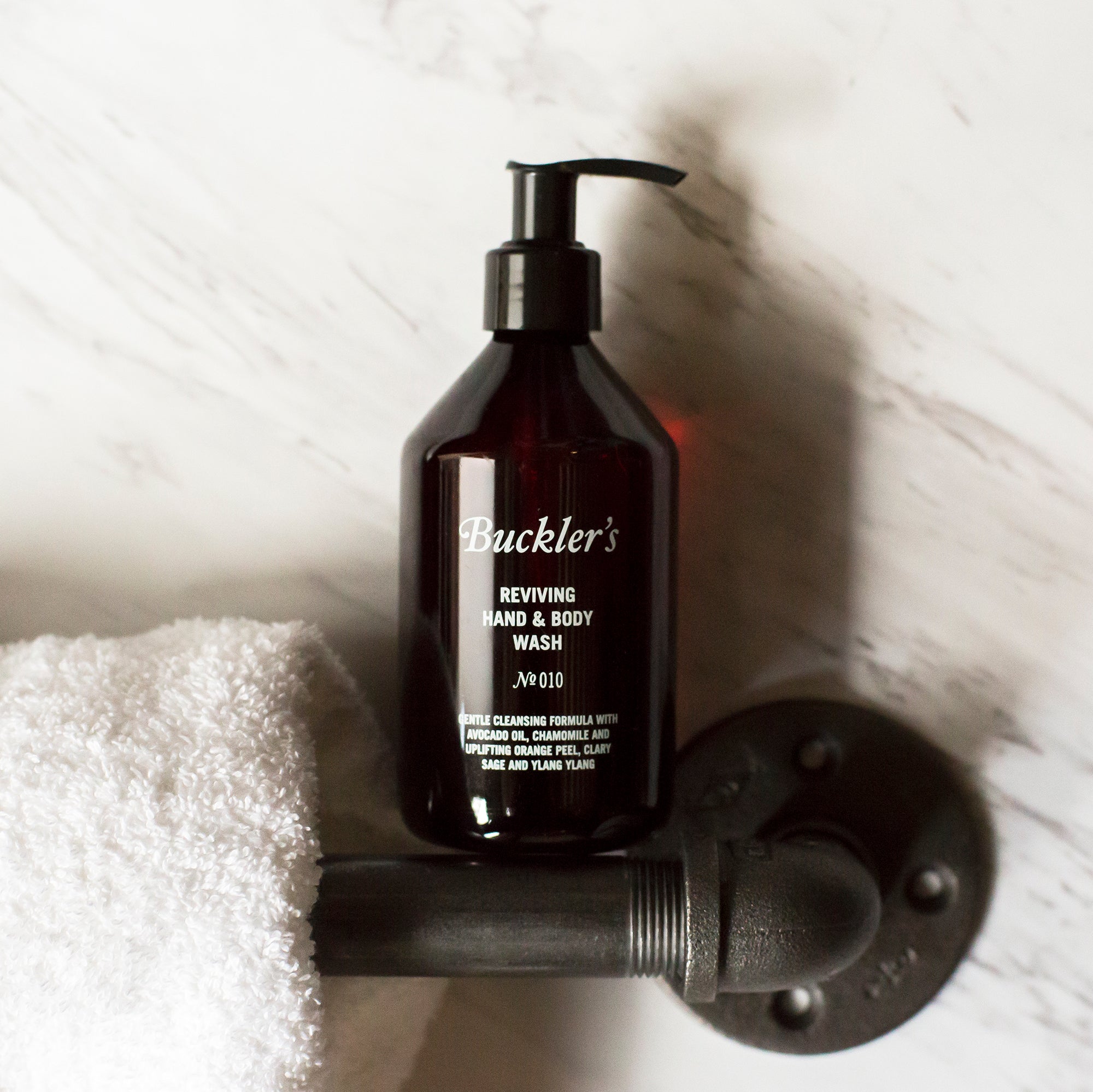 The Buckler's Reviving Hand & Body Wash in a red wine-colored bottle with a black pump top stands on a silver bathroom handle bar next to a white towel against a white and gray marble background.
