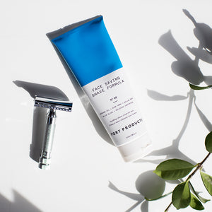 Port Products Face Saving Shave Formula white and blue tube on a white counter with a silver razor to the left, and the branch of a plant to the right.