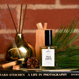The Motley Palo Santo Fragrance in a glass bottle and a black pump cap rests on a counter with pine needles, palo santo wood smoking, and a reed incense distributor behind it.