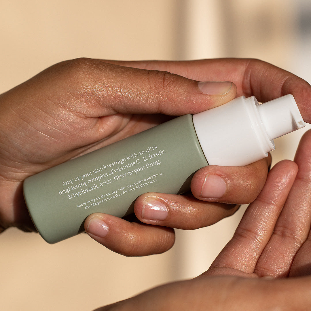 A hand holds a Glow Getter C E Serum in a sage green glass bottle with a white pump cap and is about to pump it into the other hand.