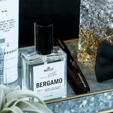 The Motley Bergamo Fragrance in a glass bottle with clear liquid and a black pump cap rests in a gray tray with gold trim next to a black bow tie, Motley Comb, Conditioner, and a fancy alcohol bottle with gold liquid.