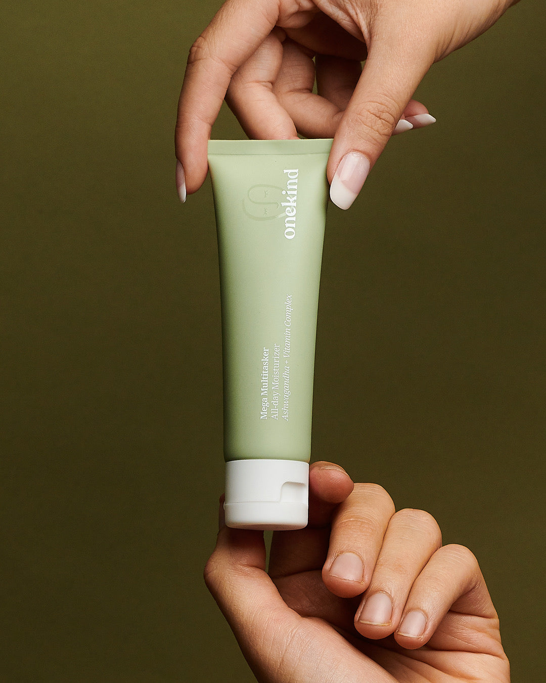 Two hands hold the Mega Multitasker All-Day Moisturizer in a sage green tube with a white cap against a forest green background.