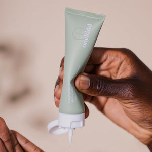 A Mega Multitasker All-Day Moisturizer in a sage green tube with a white cap that is open is being squeezed by one hand onto the palm of another, and the cream is white and velvety.