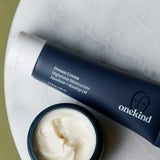 A Dream Cream Nighttime Moisturizer in a dark blue tube with a white cap on a white marble background with the cream exposed below to show the smooth, velvety cream-colored texture of the moisturizer.
