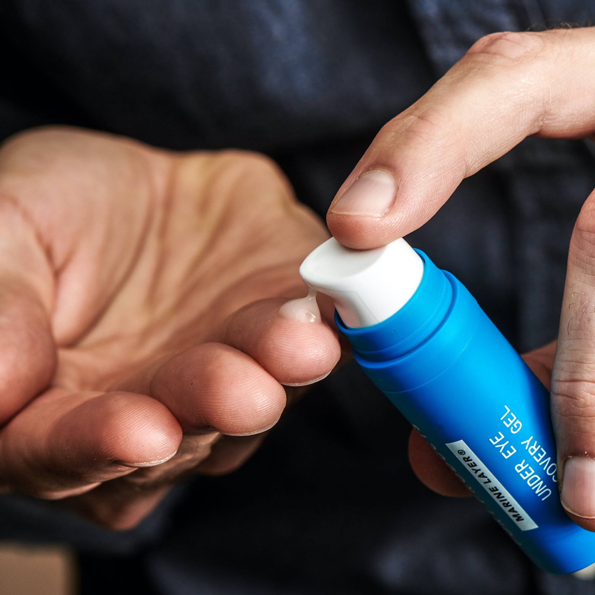 Marine Layer® Under Eye Recovery Gel blue bottle with white pump. Hands pumping liquid from bottle onto fingers