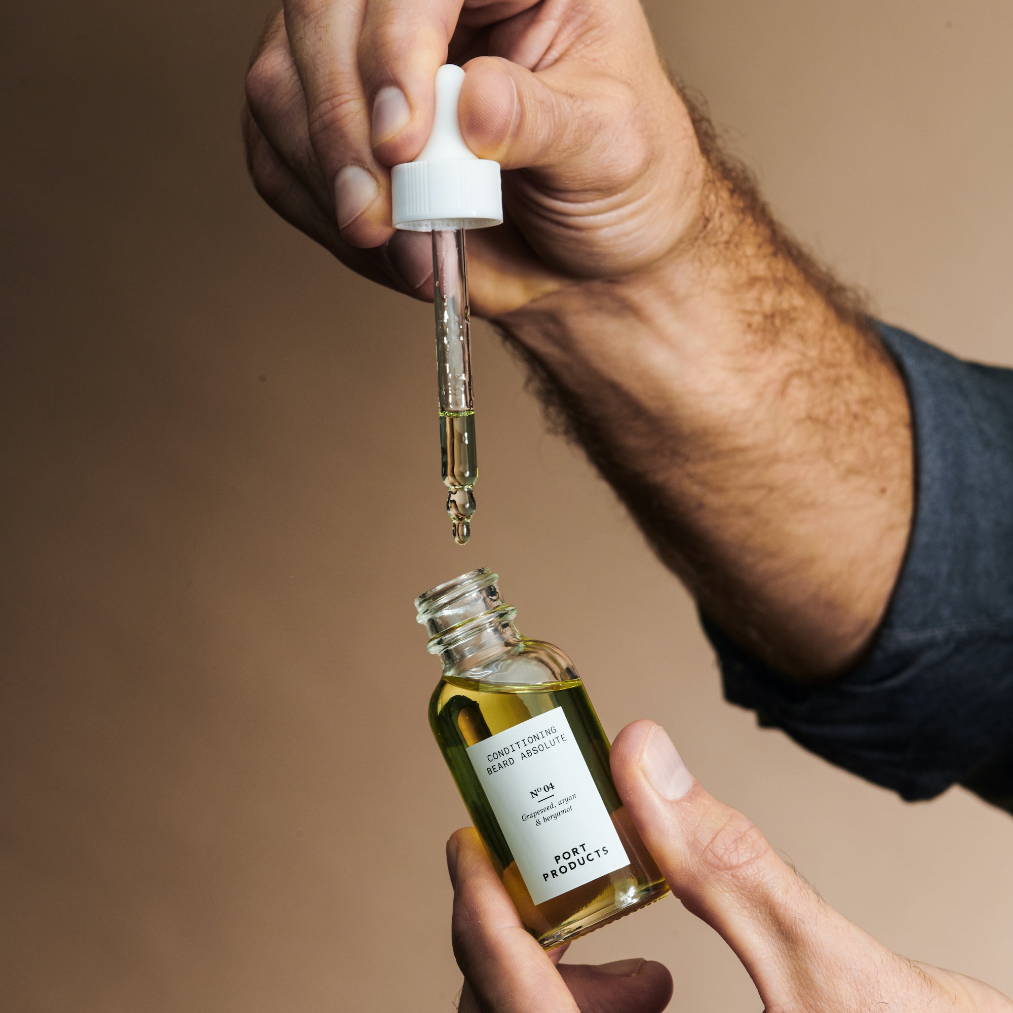 Port Products Conditioning Beard Absolute bottle and dropper in man's hands. White dropper lifted out of bottle to show gold liquid in dropper