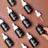 Multiple Midnight Magic PM Serums in dark blue glass jars with white dropper caps are spread on a pale maroon-colored background in a sorted pattern, and the one in the bottom right corner is open with the dropper set to the side and a hand is reaching toward it.