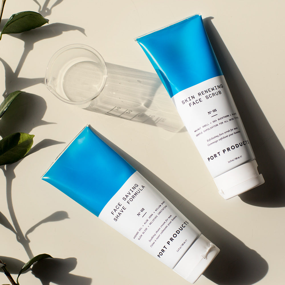 Smooth Sailing Shave Duo of shave formula and face scrub in white and blue tubes rest on cream-colored counter with a glass of water and plant leaves.