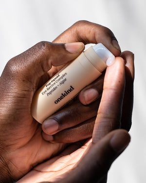 A hand holds a Pep Squad Eye Revive Complex in a light tan tube with a white pump top and is pumping the white-colored gel onto the fingertip of the other hand.