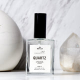 The Motley Quartz Fragrance in a clear glass bottle with clear liquid and a black pump cap sits on a white counter with a white marble background and a large, clear crystal.