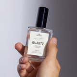 A hand holds The Motley Quartz Fragrance in a clear glass bottle with clear liquid and a black pump cap against a gray background.