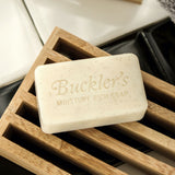 The cream-colored bar of Buckler's Moisture Rich Soap sits on a wooden grate against black and white bathroom tiles.
